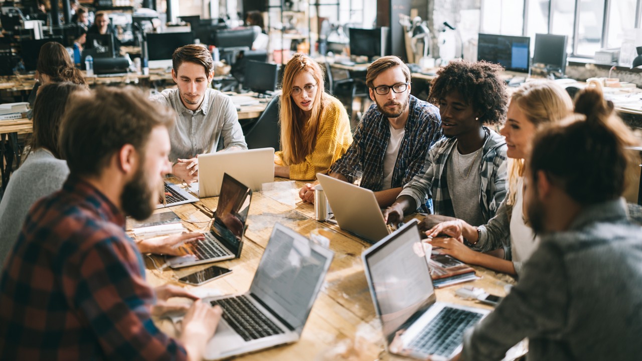 Farklı etnik kökenden web geliştiriciler, modern ofiste dizüstü ve tabletlerle web sitesi optimizasyon stratejileri üzerine çalışıyor.